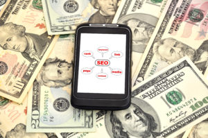 A cell phone with an SEO icon on it sitting on top of a pile of money, showcasing the expertise of a digital marketing agency in SEO.